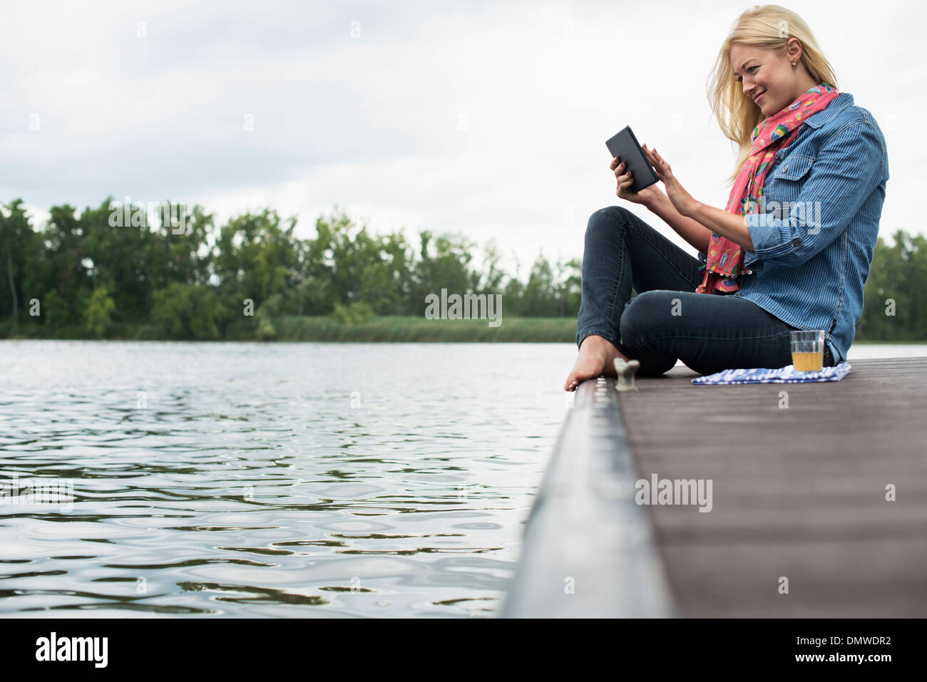 Eine Frau sitzt auf einem Steg an einem See mit einem digitalen Tablet. Stockfoto