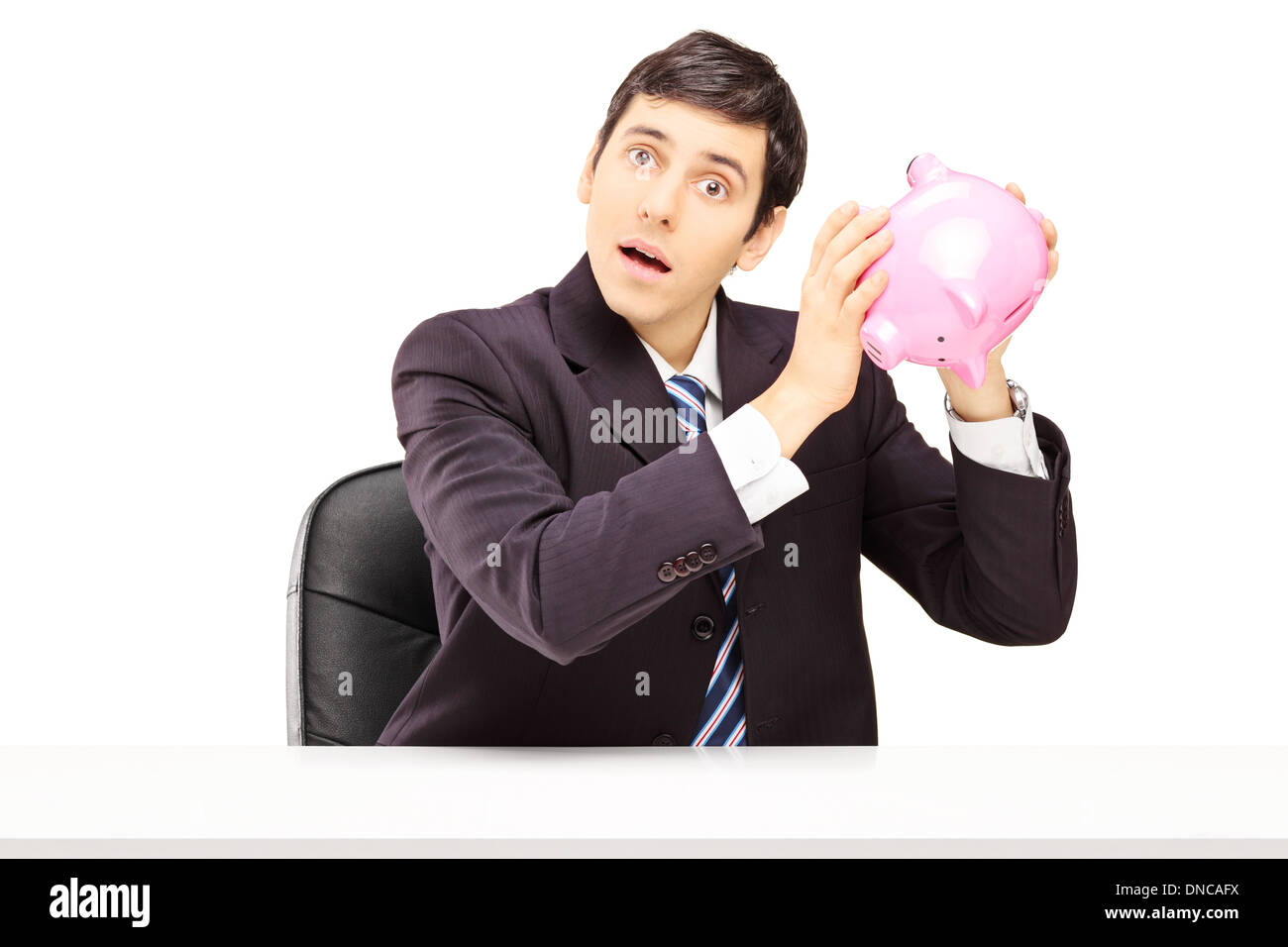Junger Geschäftsmann sitzt auf seinem Schreibtisch ein Sparschwein anhören Stockfoto