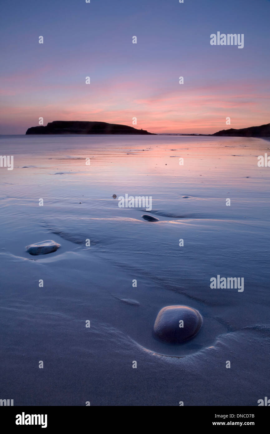 Kieselsteine und nassen Sand links von der zurückweichenden Flut am Strand von Rhossili Bay bei Sonnenuntergang. Burry Holms können in der Ferne gesehen werden. Stockfoto