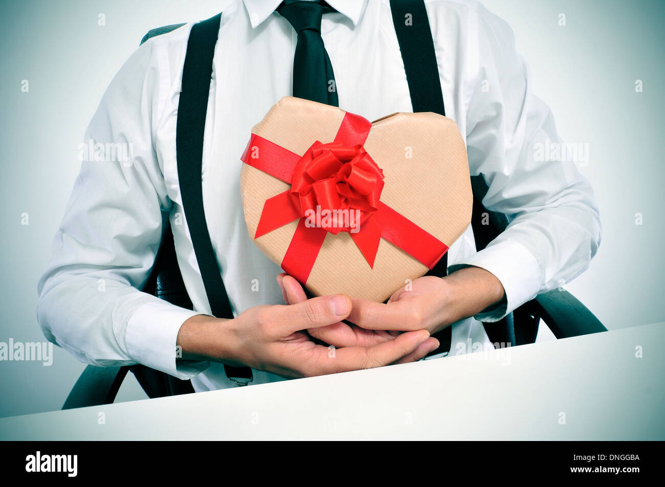 Mann in einem Anzug sitzt in einer Tabelle mit einem herzförmigen Geschenk in den Händen Stockfoto