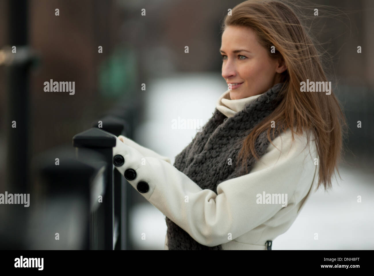 Eine schöne junge Frau in einem weißen Mantel und grauen Schal lächelt auf einer Brücke im Winter. Stockfoto