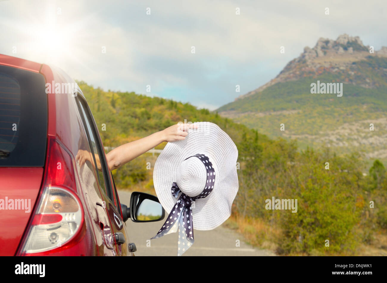 Frau zeigt Sonnenhut aus Autofenster mit Bergen im Hintergrund. Reisen-Urlaub-Konzept. Stockfoto