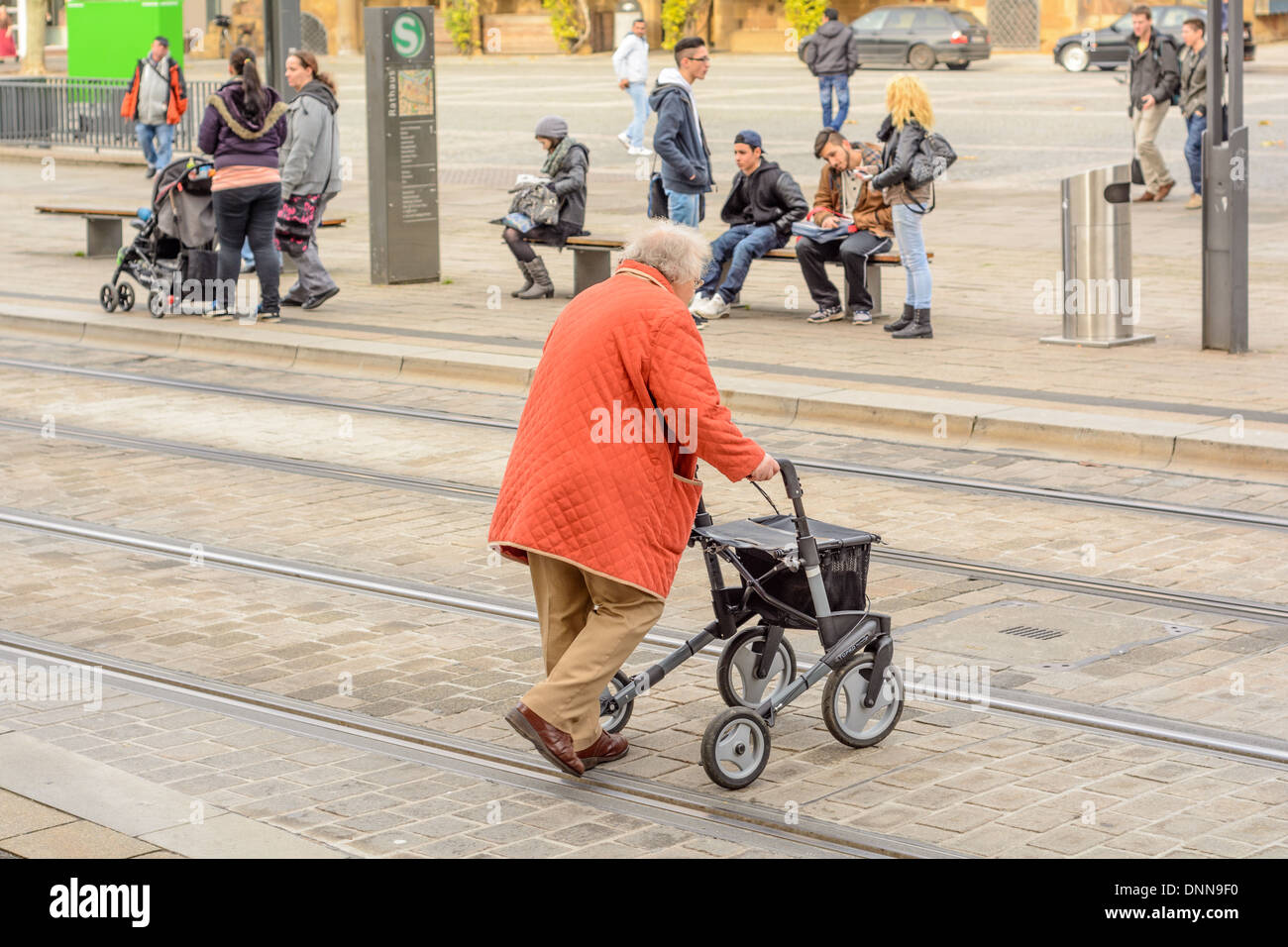 Alte Dame mit Orangen Mantel und Rollator mit der Handbremse, überqueren einer Straße mit Embeded Straßenbahnschienen – Heilbronn Deutschland Stockfoto