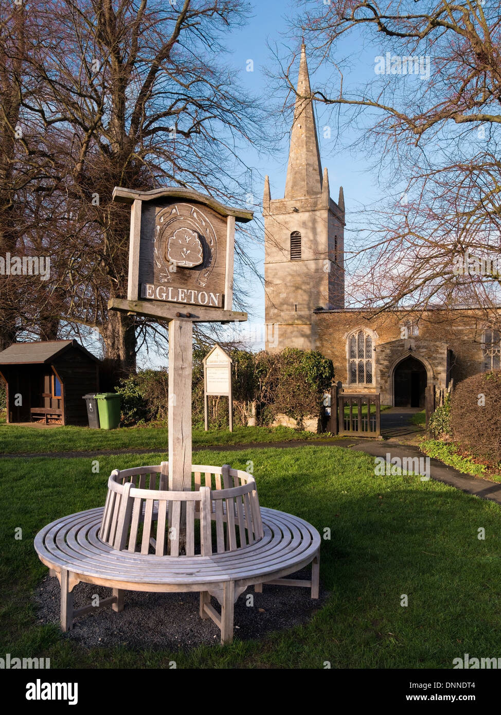 Runde Sitzfläche und Dorf Holzschild auf Egleton Dorfplatz mit Kirche St. Edmund hinaus Egelton, Rutland, England, UK. Stockfoto
