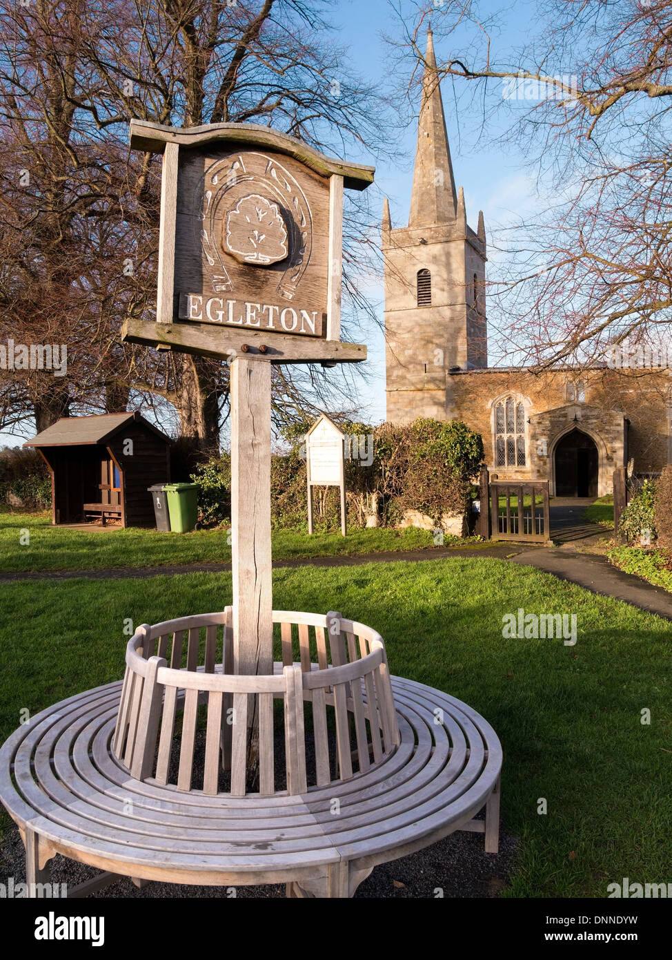 Runde Sitzfläche und Dorf Holzschild auf Egleton Dorfplatz mit Kirche St. Edmund hinaus Egelton, Rutland, England, UK. Stockfoto