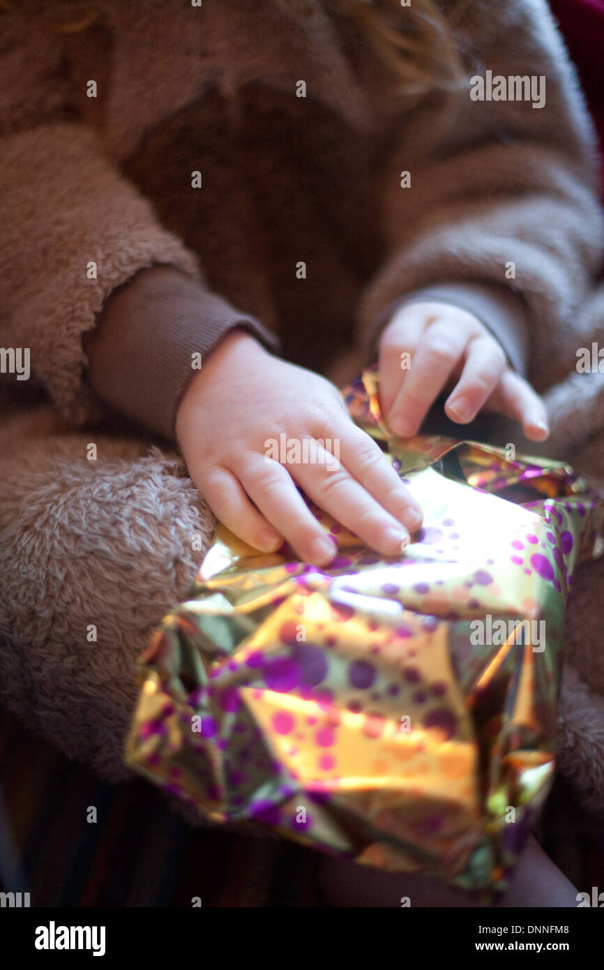 Weihnachtstag und ein kleines weibliches Kind gekleidet in ein Bär Strampler sorgfältig öffnet sich ein kleines Geschenk verpackt in metallic-Papier Stockfoto