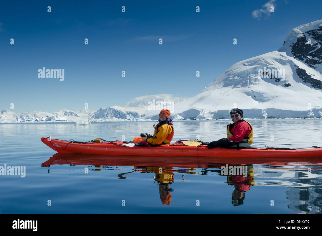 Kajakfahren Cuverville Island Antarktische Halbinsel // CUVERVILLE ISLAND, Antarktis — Kajakfahrer gleiten über die spiegelartige Oberfläche des ruhigen Wassers rund um Cuverville Island auf der Antarktischen Halbinsel unter hellem, sonnigem Himmel. Die perfekten Reflektionen schaffen eine faszinierende Symmetrie zwischen den Kajakfahrern, der eisigen Landschaft und dem klaren blauen Himmel und bieten ein seltenes und ruhiges antarktisches Paddelerlebnis. Stockfoto