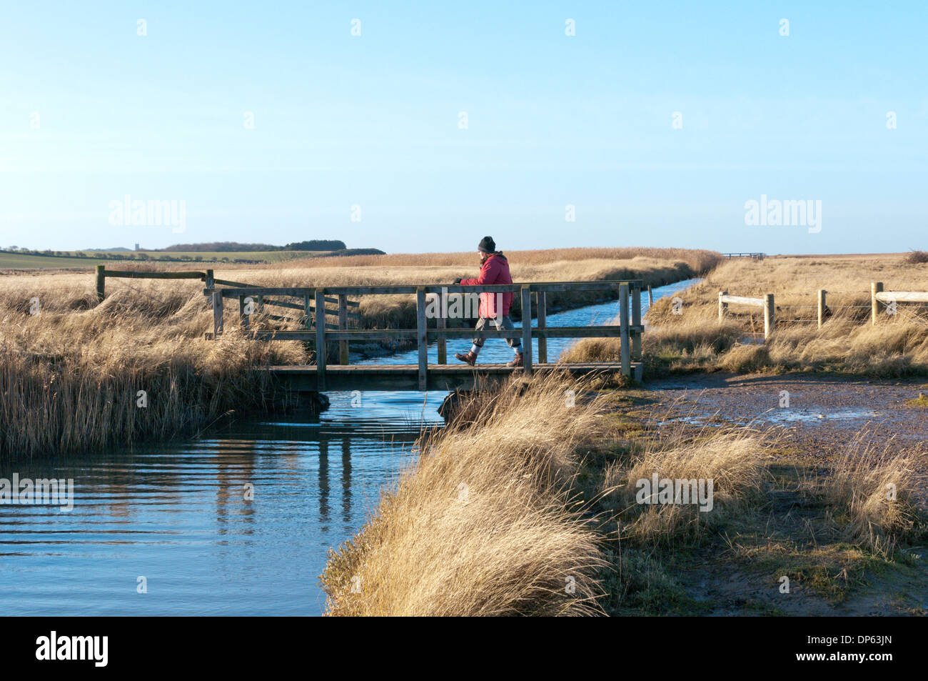 Eine Frau, die über einen kleinen Steg auf Salthouse Marshes in der Nähe von Cley an der North Norfolk-Küste. Stockfoto