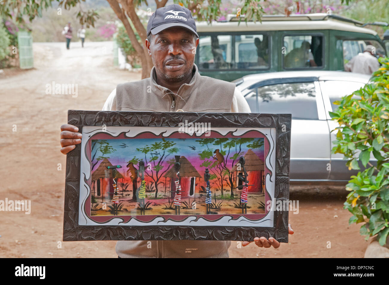 Mann zeigt afrikanische Malerei in geschnitzten Holzrahmen Darstellung Dorf Rondavel Hütten und Frauen zum Verkauf bei Duka Shop Kenia Stockfoto