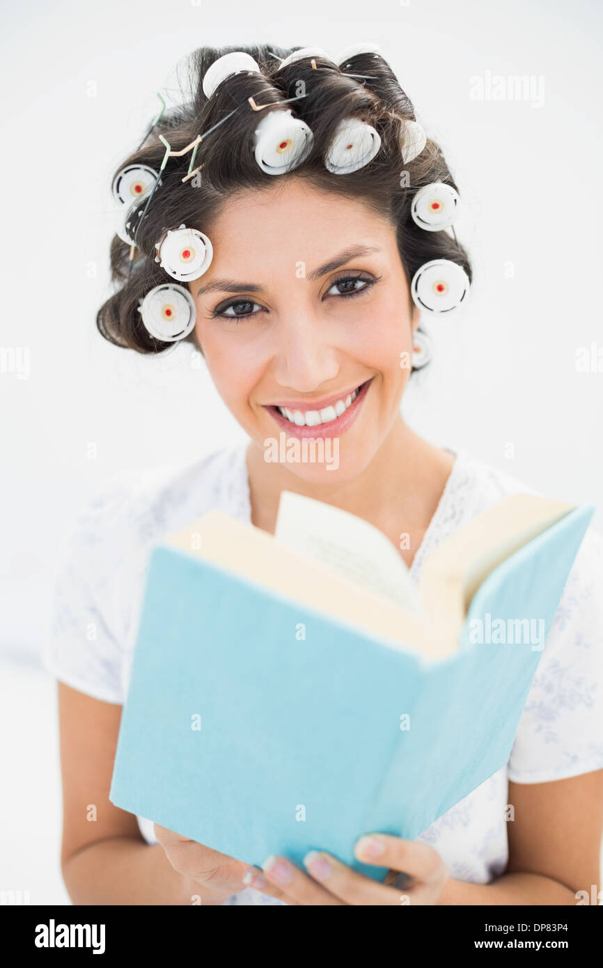 Lächelnde Brünette in Lockenwickler halten ein Buch über Bett Stockfoto