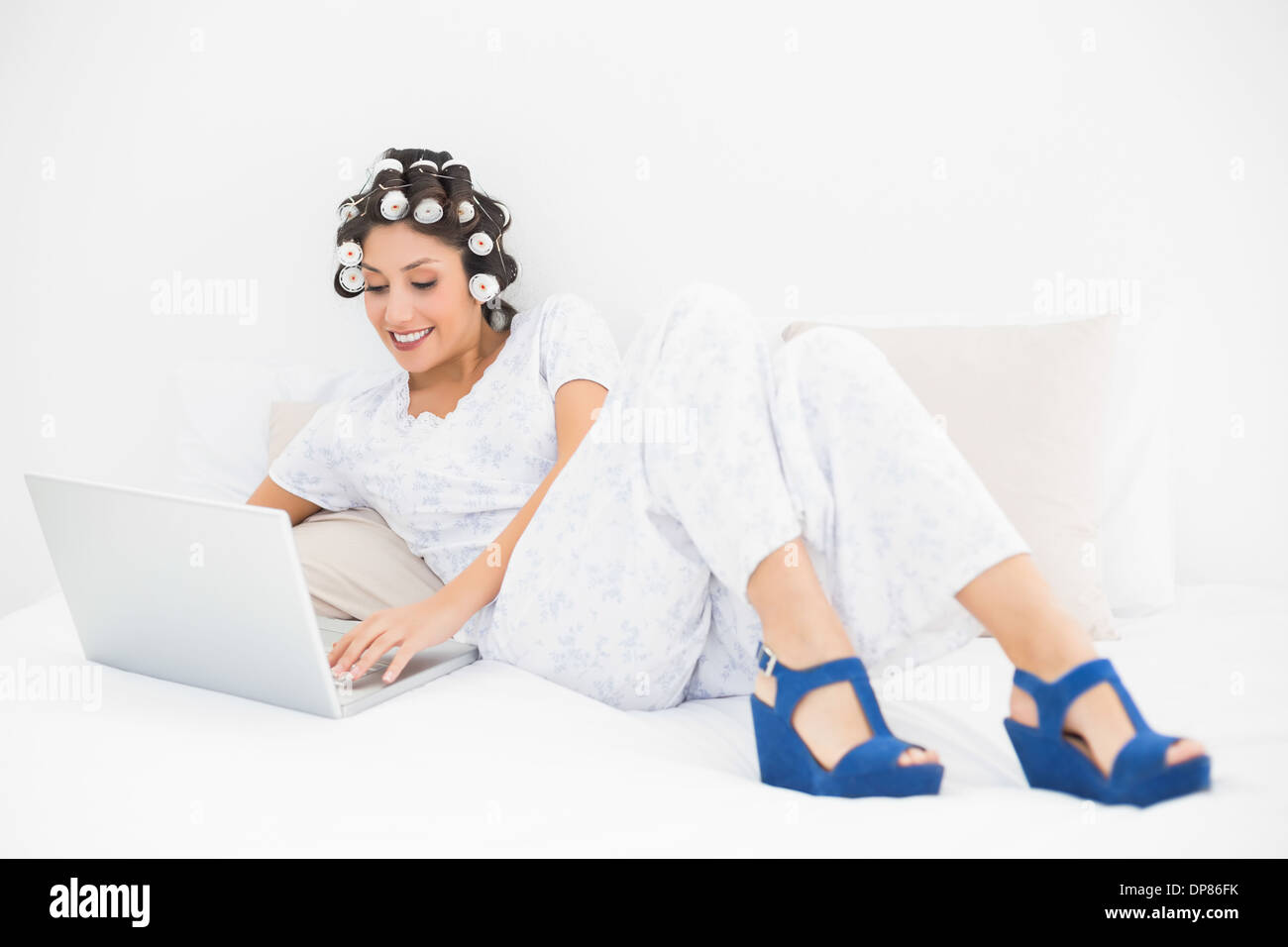 Lächelnde Brünette in Lockenwickler und Keil Schuhe mit ihrem Laptop auf Bett Stockfoto