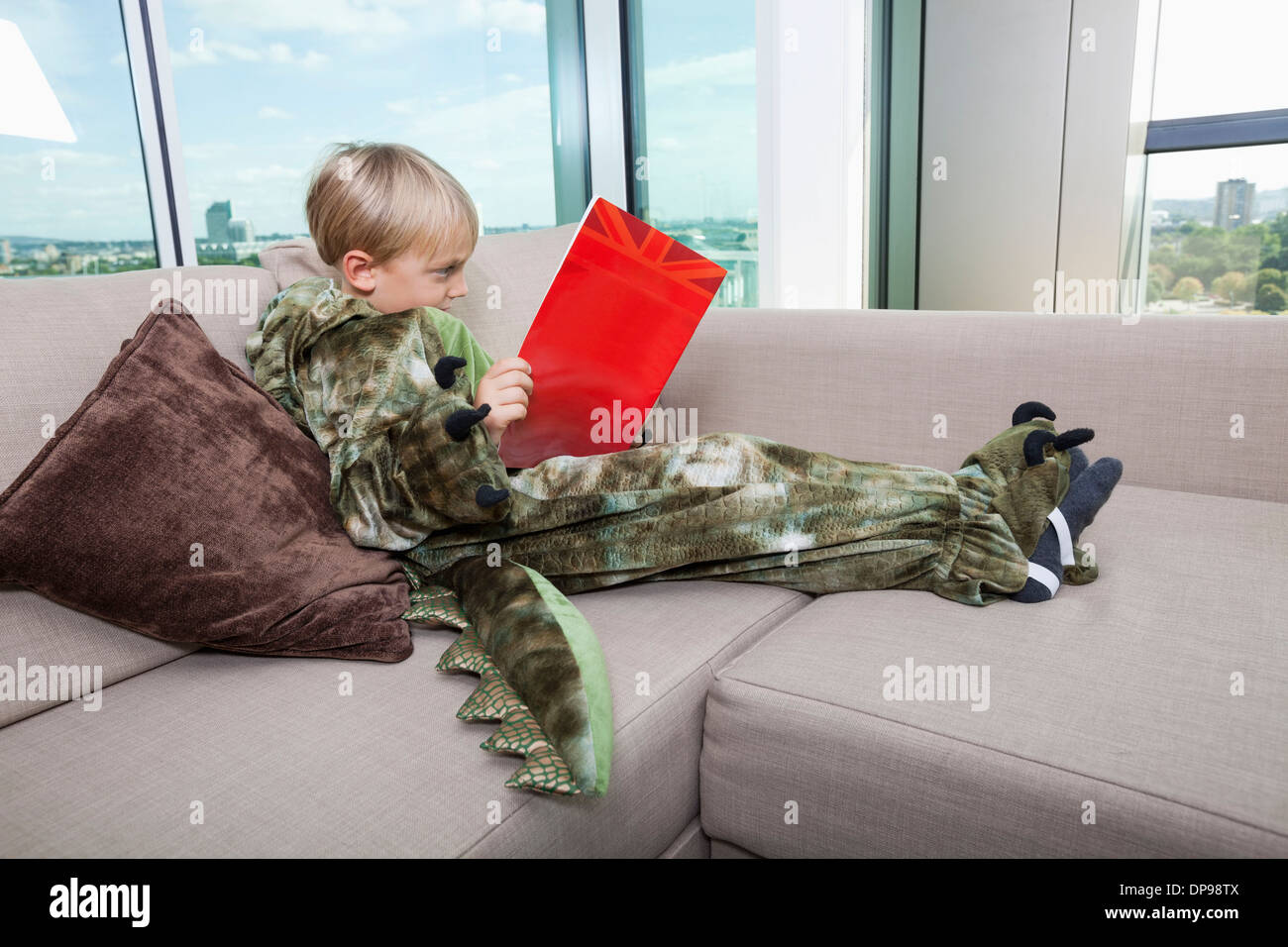 Junge im Dinosaurier Kostüm Märchenbuch auf Sofa zu Hause lesen Stockfoto