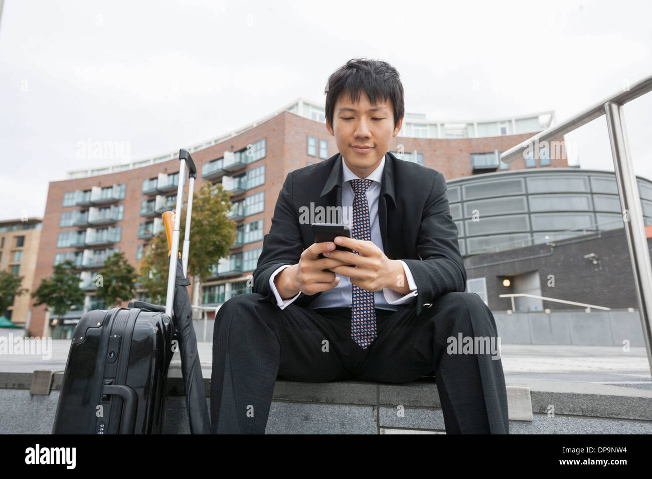 Asiatische Geschäftsmann mit Gepäck von SMS-Nachrichten über Handy gegen Gebäude Stockfoto