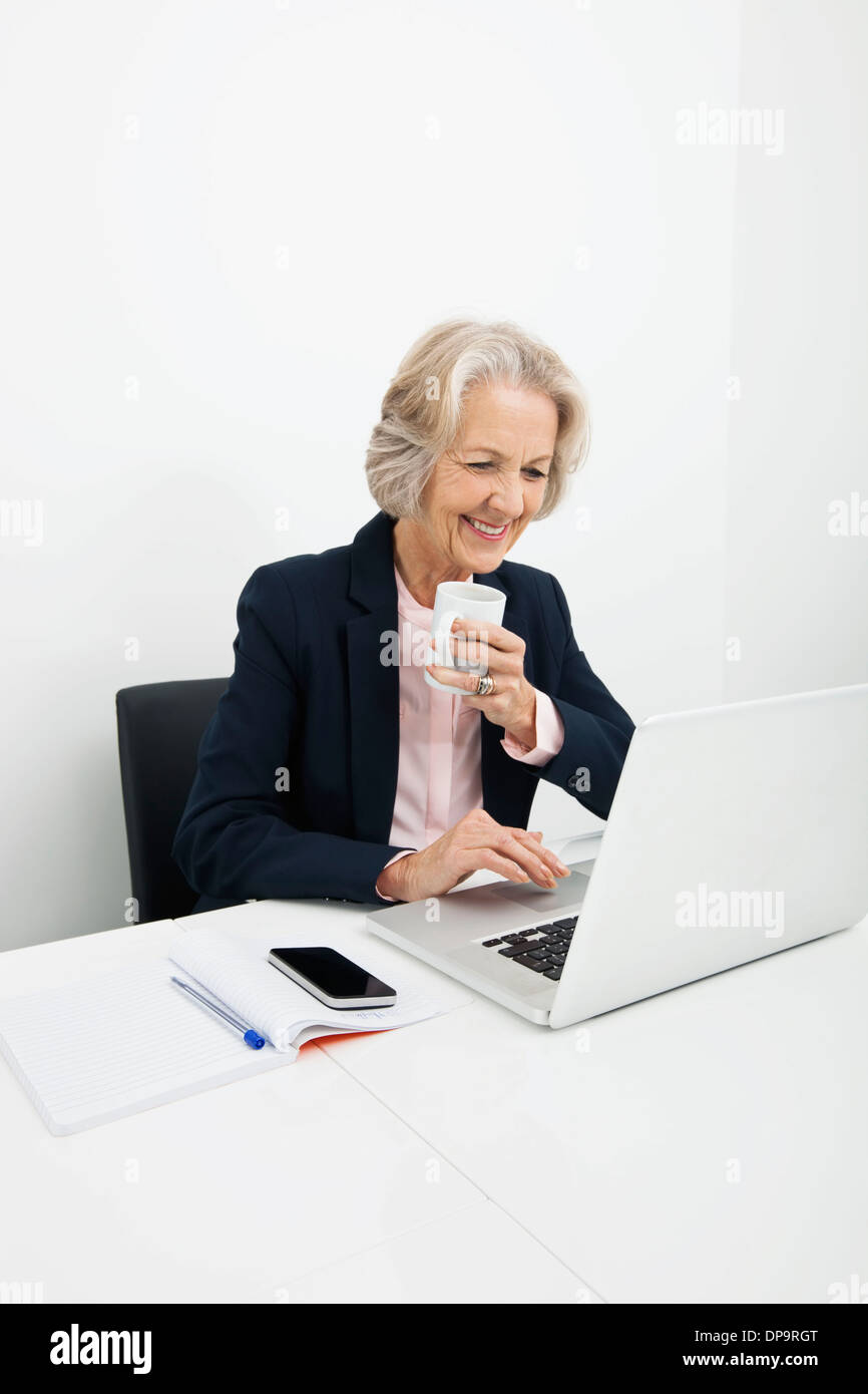 Lächelnde senior Geschäftsfrau Kaffeetrinken während mit Laptop am Schreibtisch im Büro Stockfoto