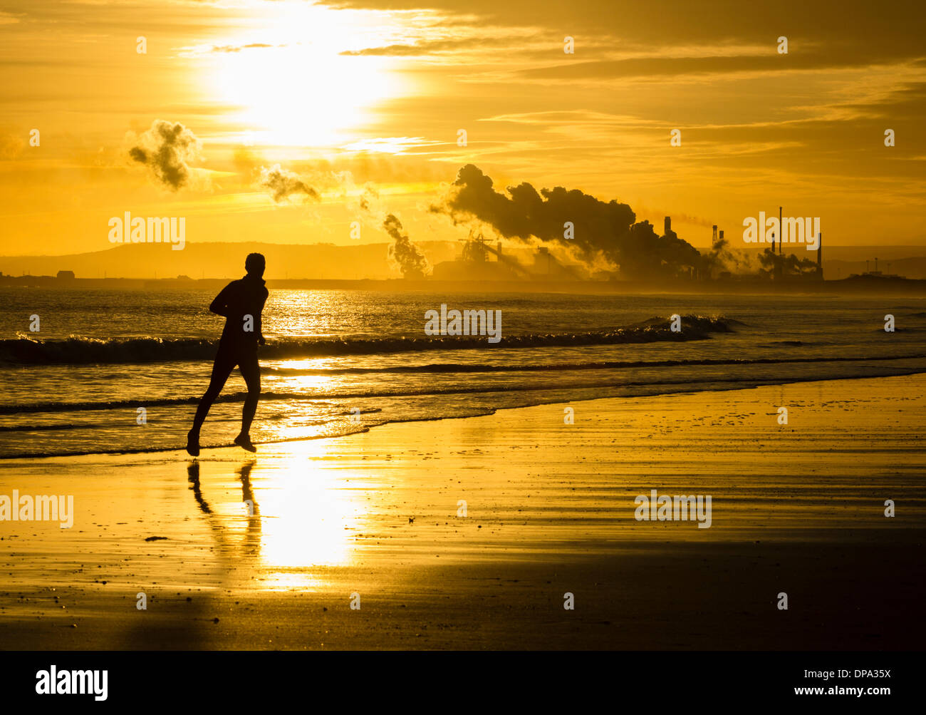 Mann, der bei Sonnenaufgang am North Gare Beach läuft.Redcar Steelworks in on the South side of the Tees Mündung, Teesmouth im Hintergrund. England. VEREINIGTES KÖNIGREICH Stockfoto