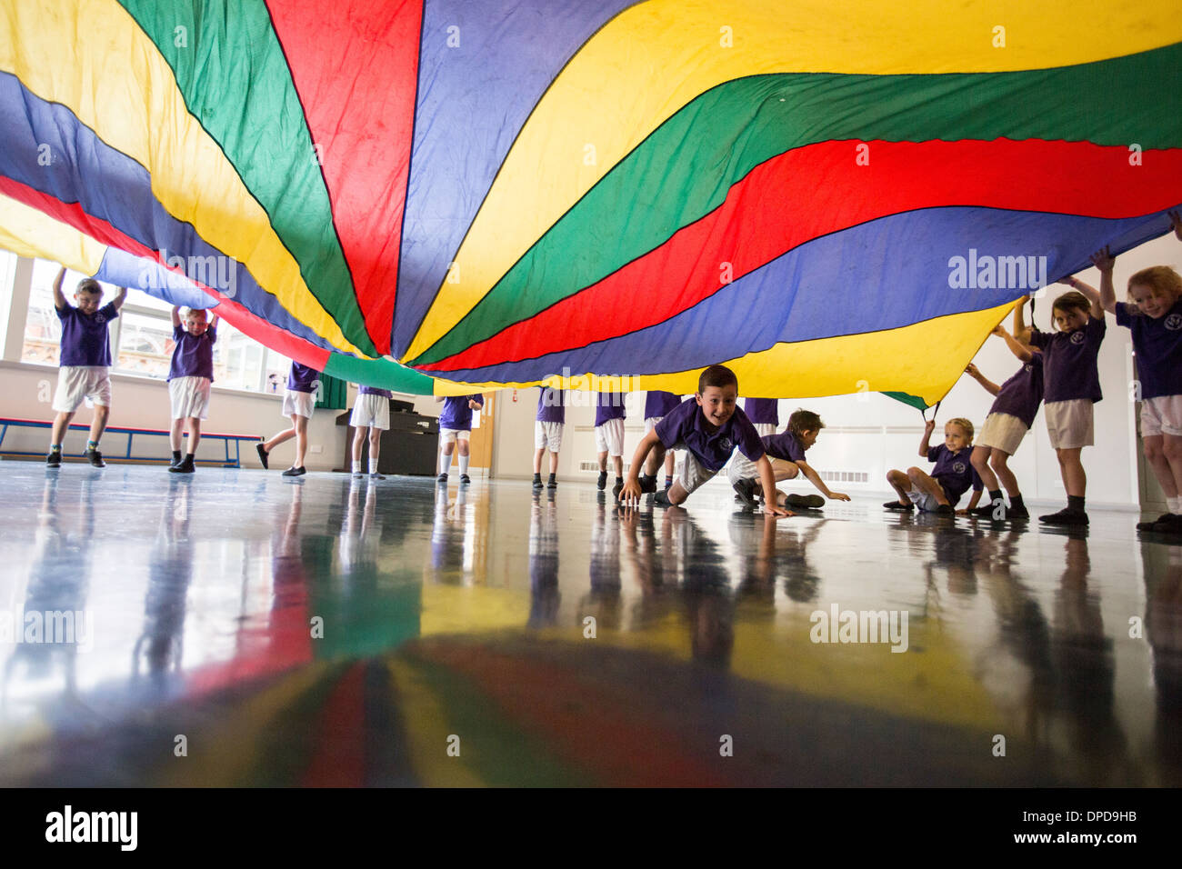 Schülerinnen und Schüler an einer UK-Grundschule spielen mit einem Fallschirm in der Aula Stockfoto
