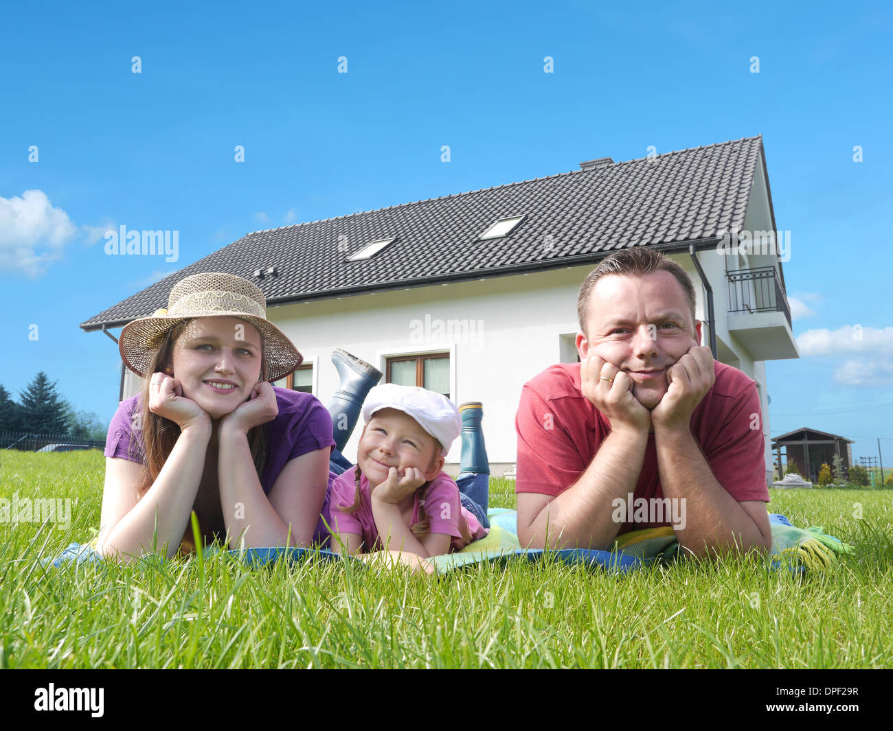 Junge Eltern mit ihren niedlichen kleinen Mädchen liegen entspannt auf dem Rasen vor dem Haus Stockfoto