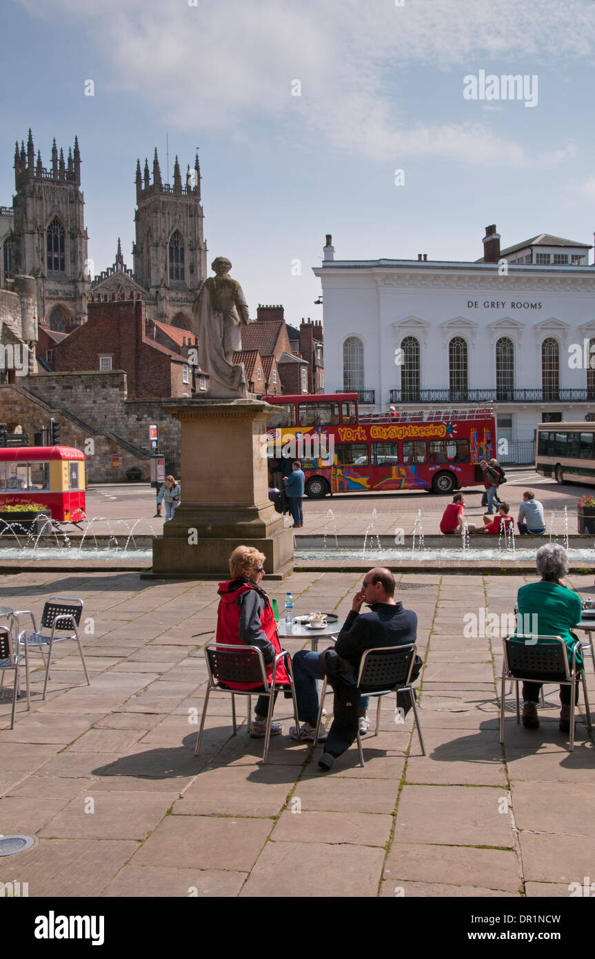 Menschen sitzen & Genießen im Freien Snack im sonnigen Piazza von Springbrunnen und Statue, Türme von York Minster & De Grau Zimmer darüber hinaus - York, England, UK. Stockfoto