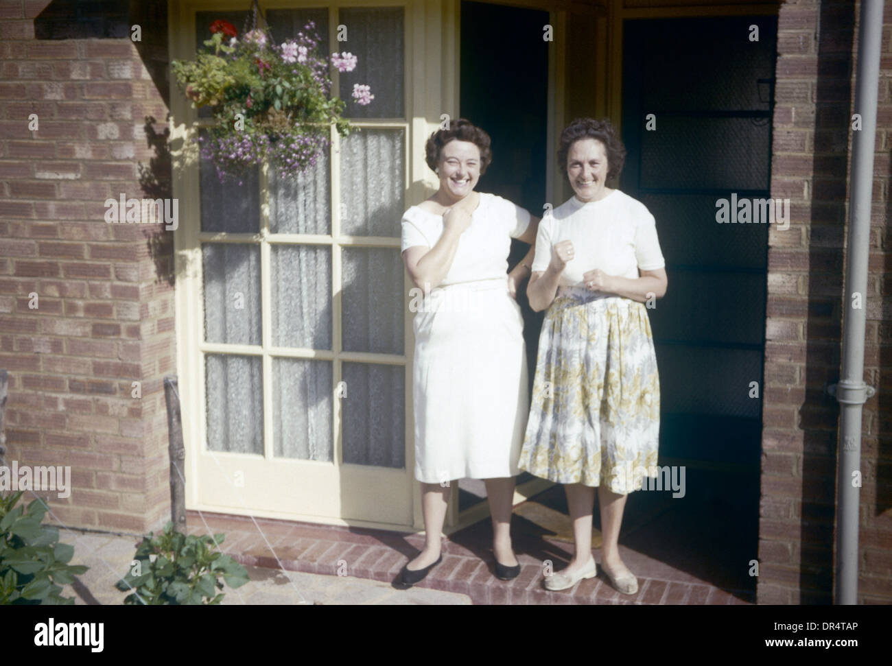 Im Sonnenschein auf der Veranda des Hauses stehen zwei 1960er Jahre Hausfrauen und Mütter. Stockfoto