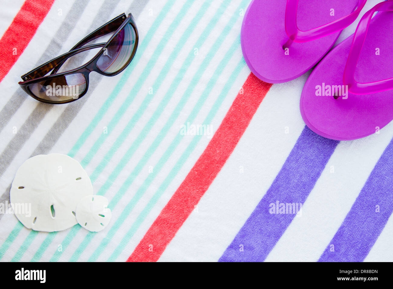 Strand-Szene mit lila Flip flops, Sand-Dollars und Sonnenbrillen auf einem gestreiften Strandtuch. Stockfoto
