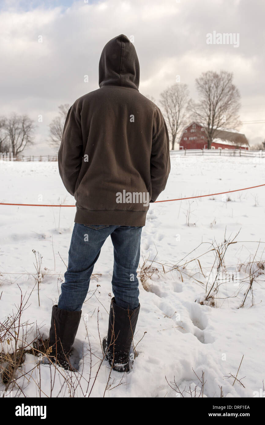 Rückseite einer Kapuze teenboy mit Blick auf einem schneebedeckten Feld gegenüber ein ländlich geprägtes Land-Bauernhof mit roten Scheune auf einem Hügel in der distanc Stockfoto