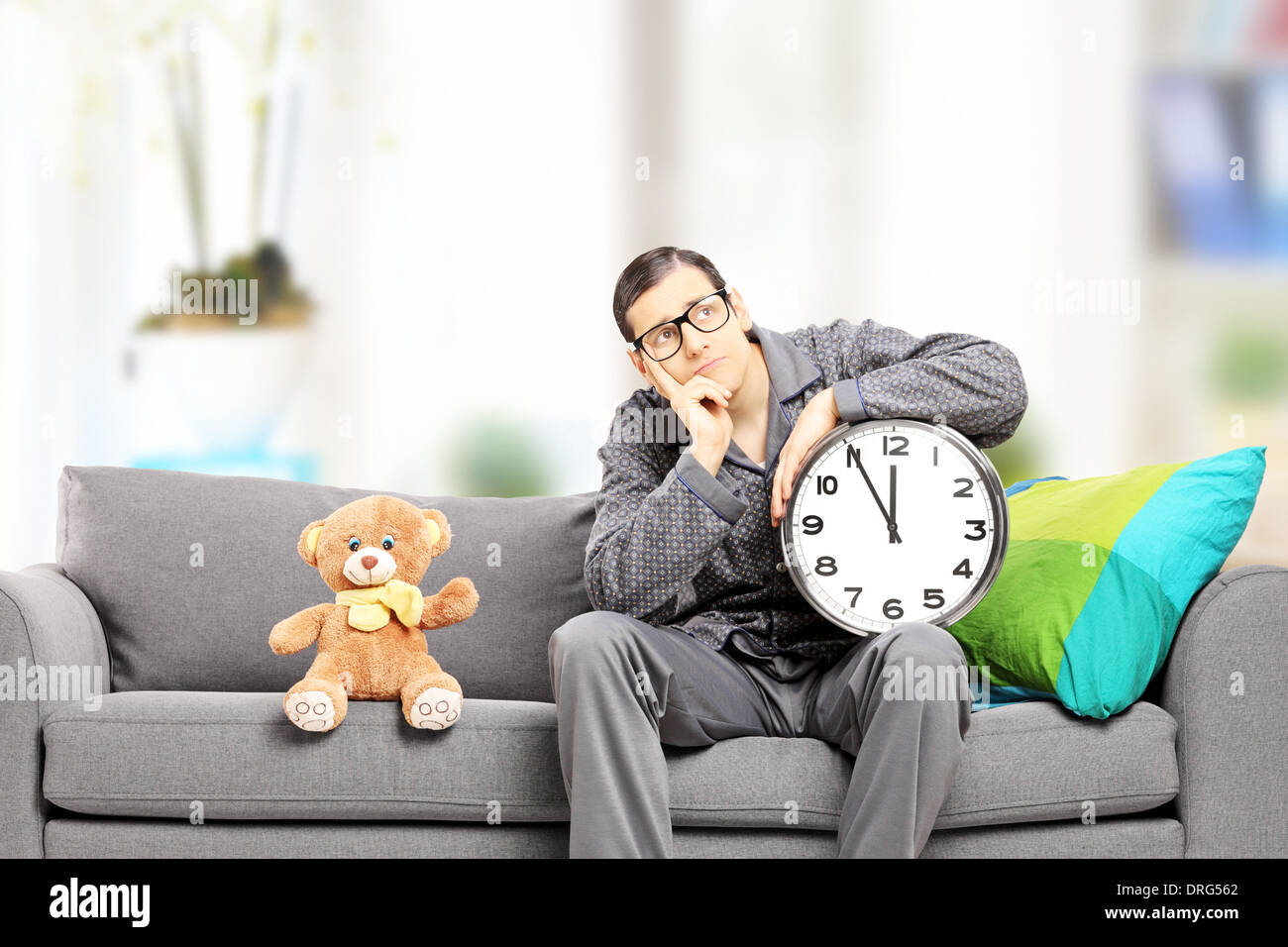 Junger Mann im Pyjama hält eine große Wanduhr, sitzen auf der Couch mit Teddybär neben ihm, zu Hause Stockfoto