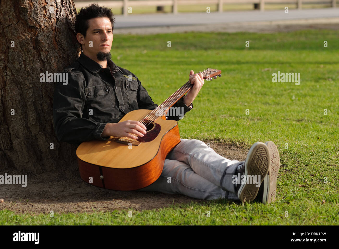Ein weißer junge Mann sitzen, lehnte sich gegen einen Baum, die Gitarre in einem park Stockfoto