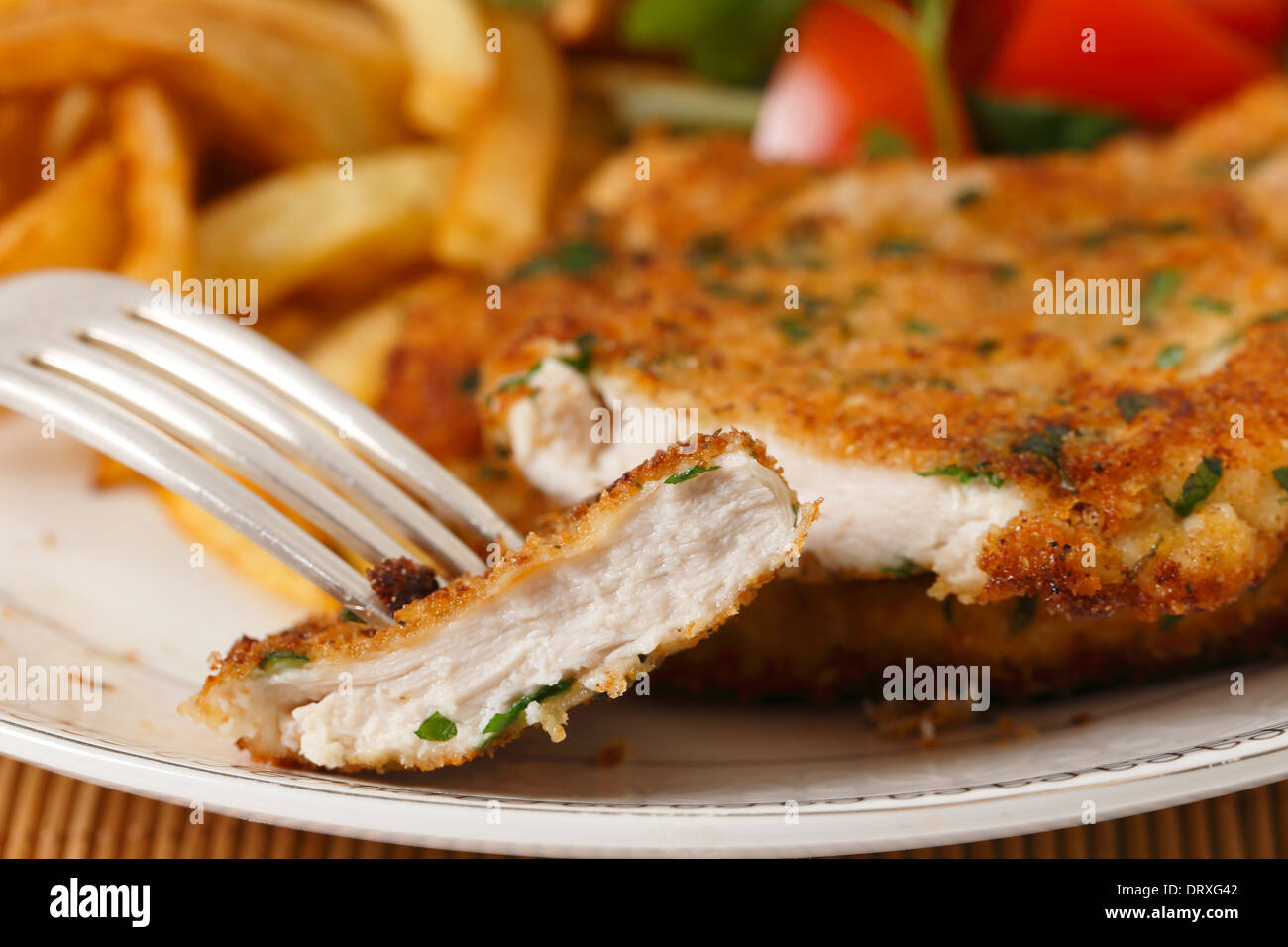 Ein Stück hausgemachte panierte Hähnchenschnitzel oder Schnitzel auf einer Gabel mit Pommes Frites und eine Tomate und grünem Salat hinter Stockfoto