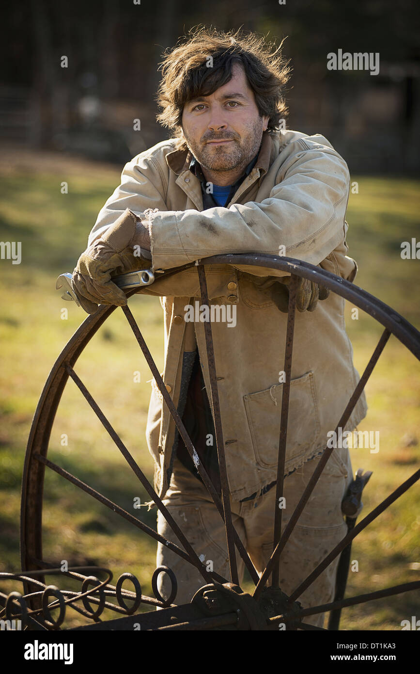 Arbeiten auf einem Biobauernhof ein Mann im Overall tragen von Arbeitshandschuhen stützte sich auf eine Runde Metall Rad mit Speichen Stockfoto