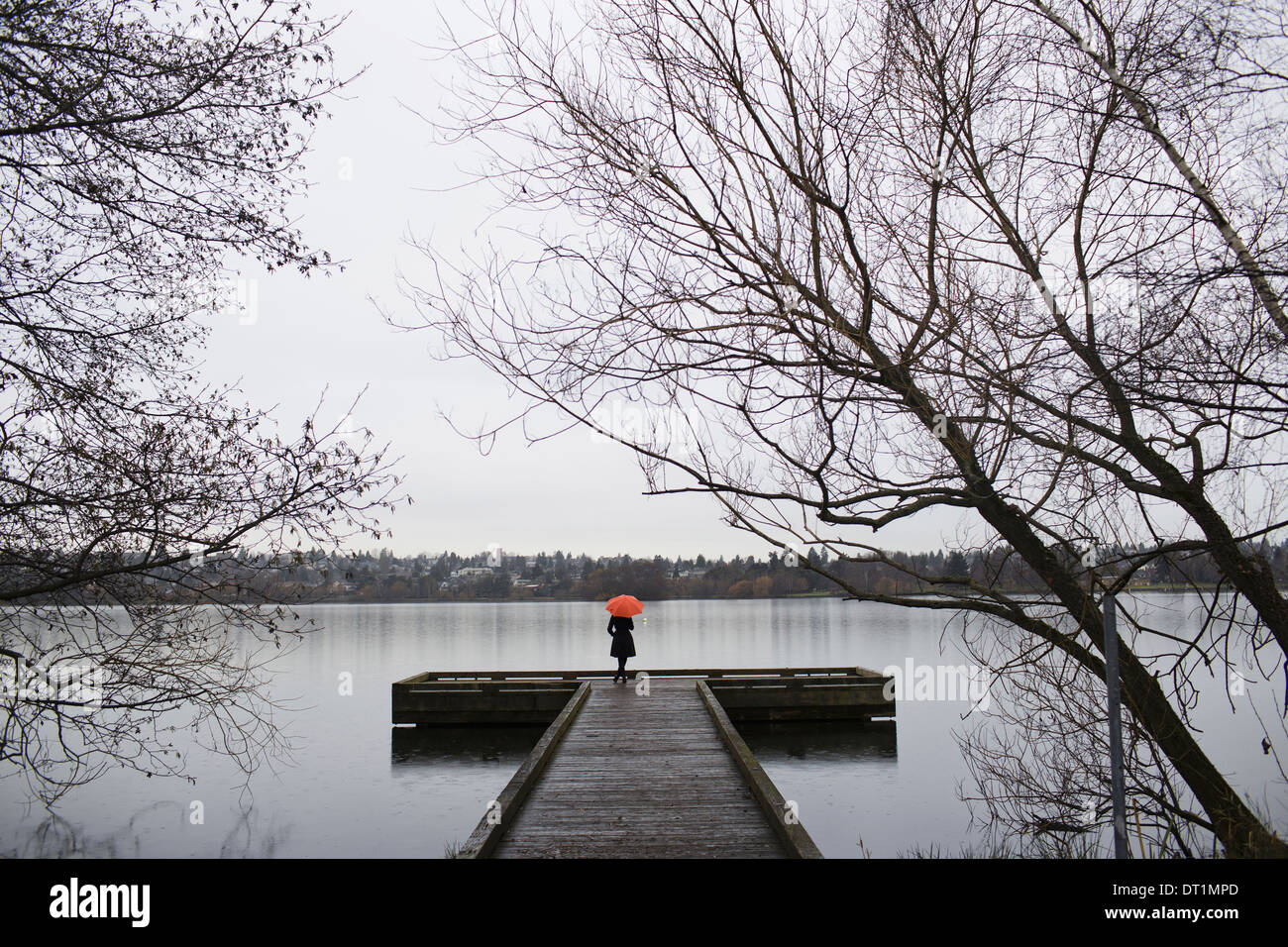 Eine Frau stand am Ende ein Dock mit einem orange Regenschirm an einem bewölkten grauen Tag in Seattle Stockfoto