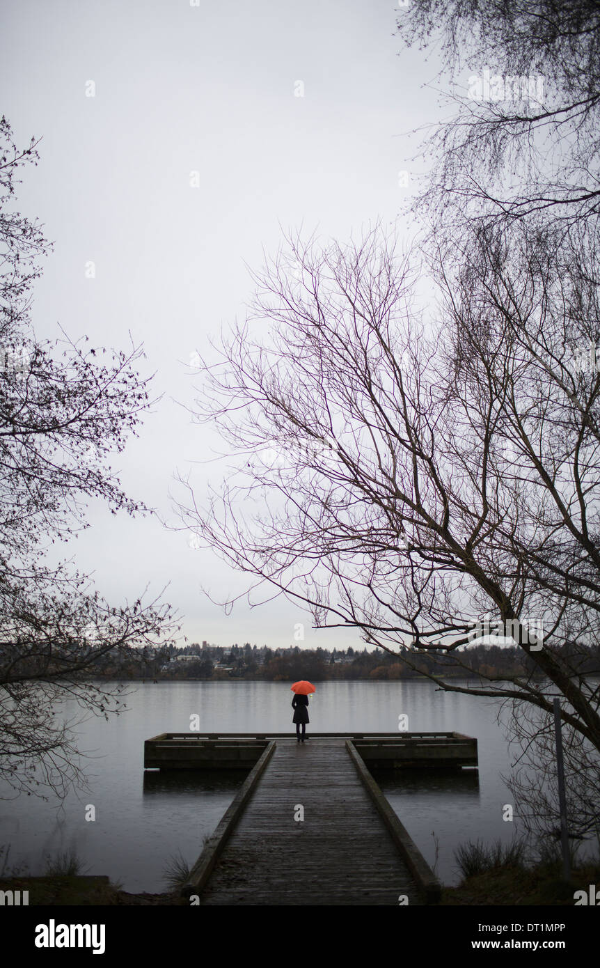 Eine Frau stand am Ende ein Dock mit einem orange Regenschirm an einem bewölkten grauen Tag in Seattle Stockfoto