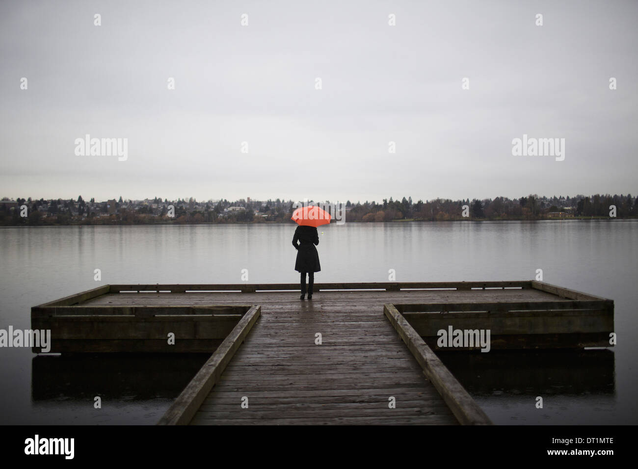 Steht eine Frau trägt alle schwarz und orangenen Regenschirm hält am Rande eines Docks auf eine graue und trübe Tag in Seattle WA Stockfoto