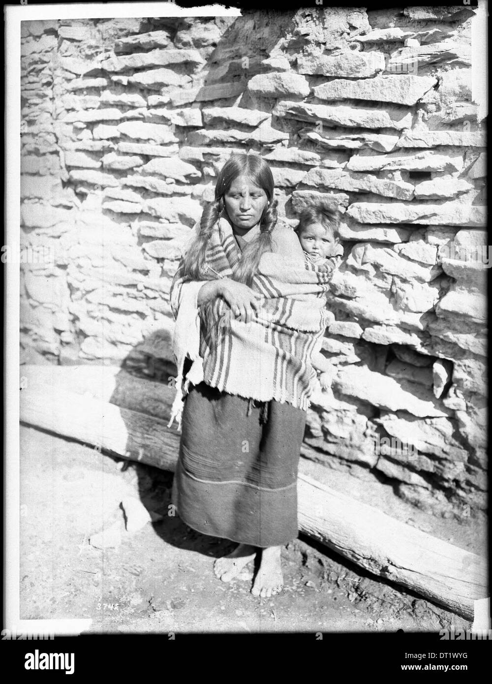 Eine Hopi-Indianermutter, die ihr Baby auf dem Rücken vor einer Steinwohnung trug, aufgenommen um 1900. Das Foto zeigt das traditionelle Familienleben und die Wohnhäuser der Ureinwohner. Stockfoto