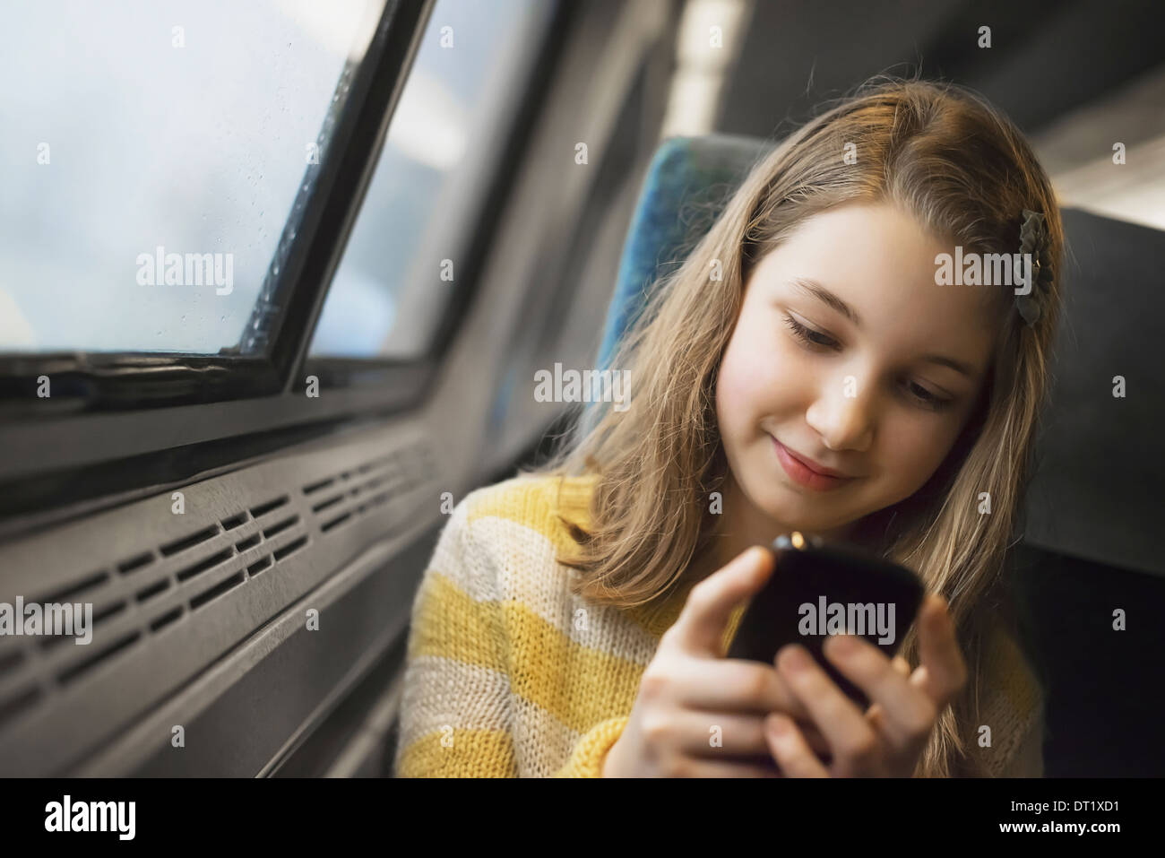 Ein junges Mädchen mit langen blonden Haaren sitzen durch ein Fenster in einem Zug mit ihrem Handy SMS und senden von Nachrichten Stockfoto