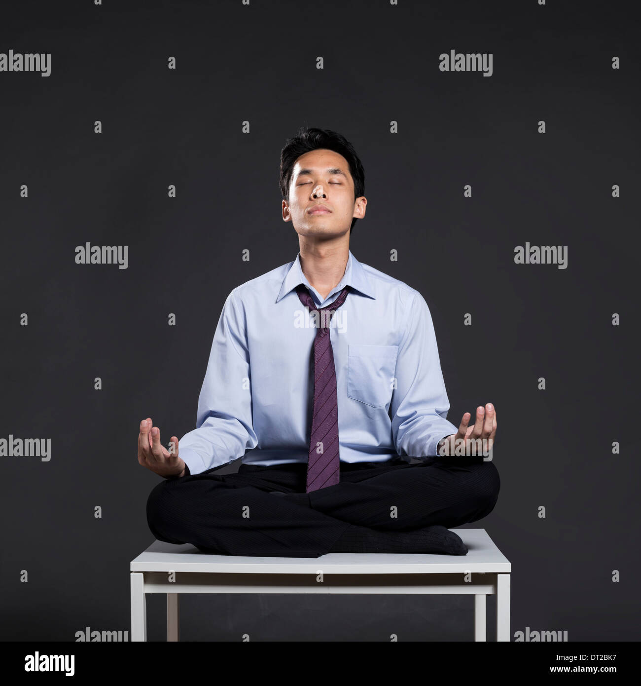 Asiatische Geschäftsmann entspannend auf Schreibtisch in Yoga Lotus-Position. Dunkel grau hinterlegt Stockfoto