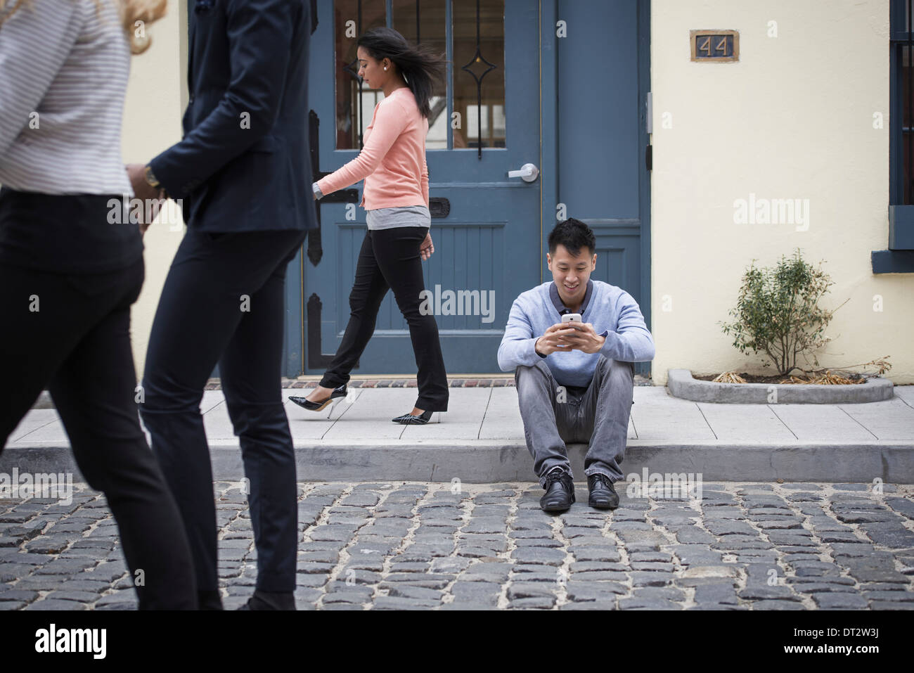 Junge Menschen im Freien auf den Straßen der Stadt im Frühling A Mann sitzt auf dem Boden Überprüfung sein Telefon und drei Passanten Stockfoto