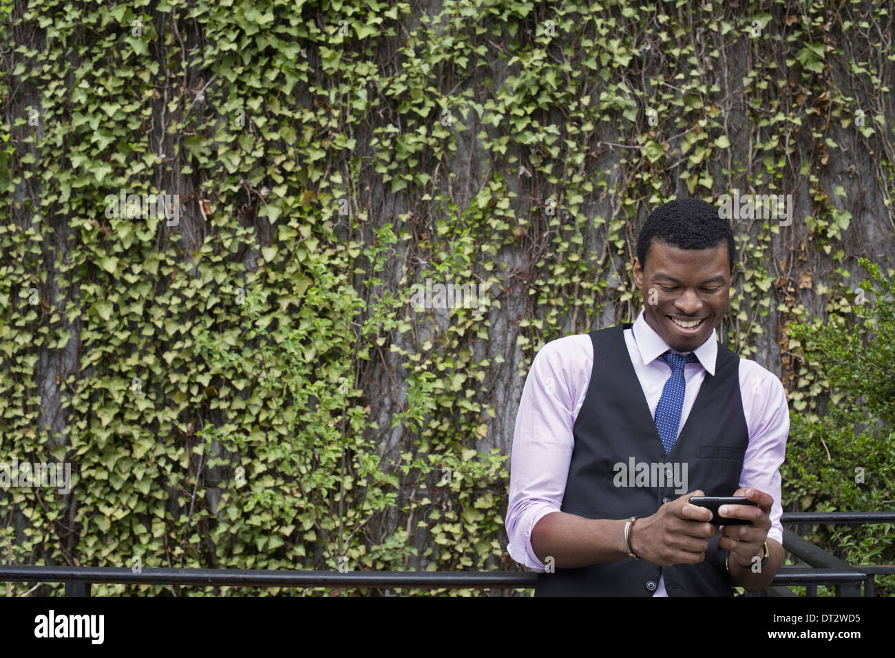 Stadtmauer Park bedeckt Kletterpflanzen und Efeu A junger Mann in eine Weste Hemd und Krawatte sein Telefon überprüfen Stockfoto