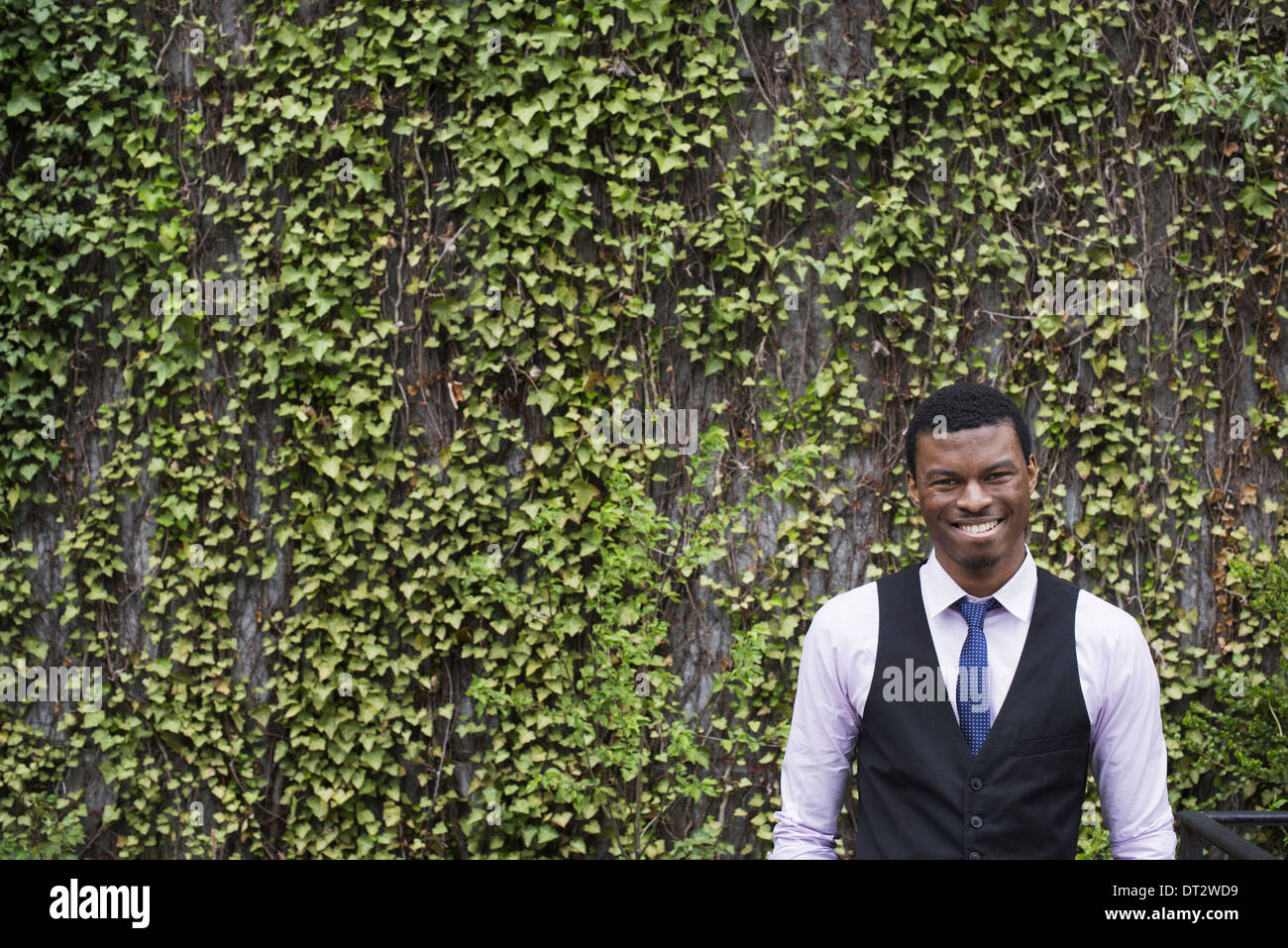 Stadtpark mit einer Wand mit Kletterpflanzen begrünt und Efeu A junger Mann in einem Weste Hemd und Krawatte Looking in die Kamera Stockfoto