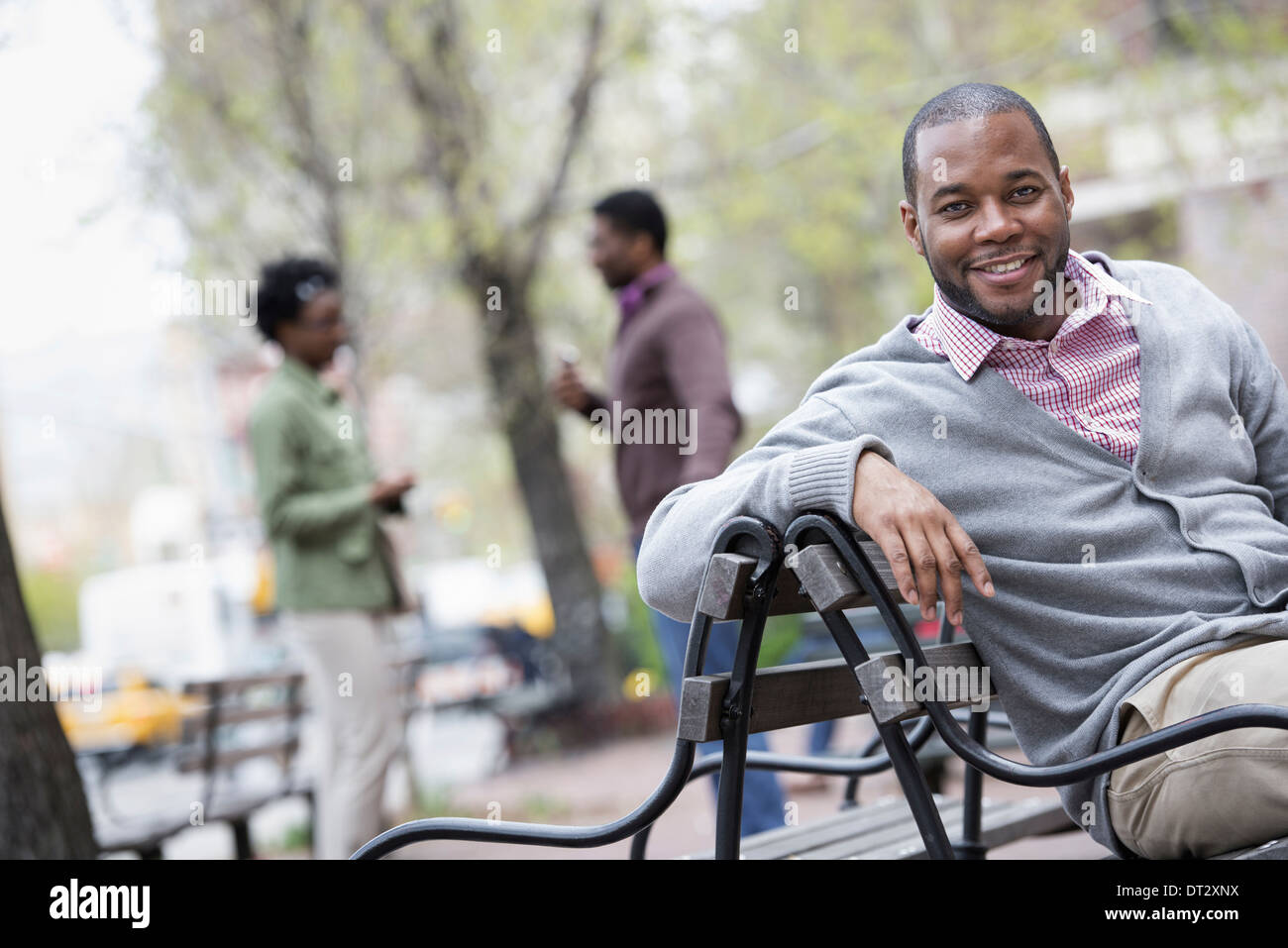 Ein Mann sitzt auf einer Bank mit zwei Personen im Hintergrund Stockfoto