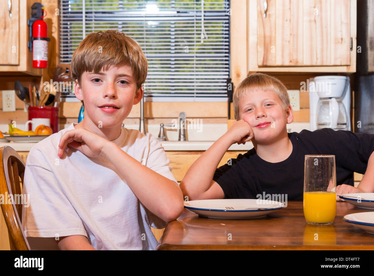2 männliche Kinder Blick in die Kamera beim Sitzen am Küchentisch mit Platten und einem Glas Orangensaft Stockfoto