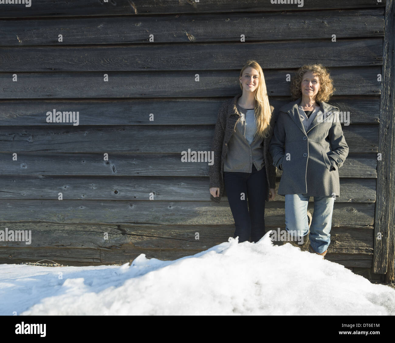 Eine Mutter und Tochter stehen an die Holzwand einer Scheune auf dem Bauernhof gelehnt. Schnee aufwärts zu ihren Füßen. Stockfoto