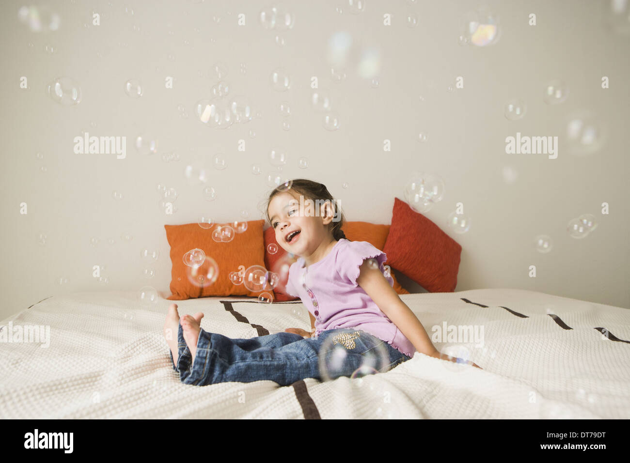 Ein junges Mädchen mit braunen Augen und dunklen Haaren in Trauben sitzen auf einem Bett vor Lachen. Seifenblasen in der Luft. Stockfoto