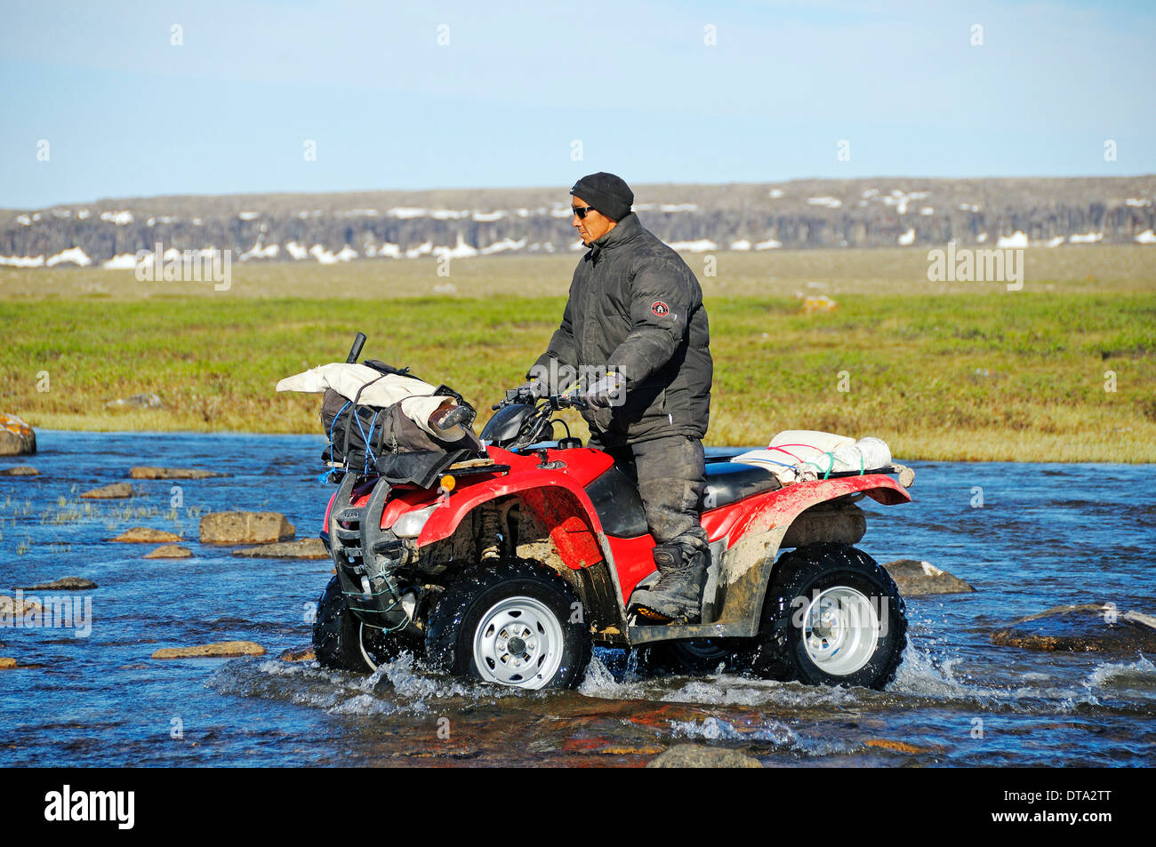 Mann von den Inuit, die Fahrrad fahren Quad, ATV, durch einen Fluss in der Tundra auf Victoria Island, früher Holman Island Stockfoto