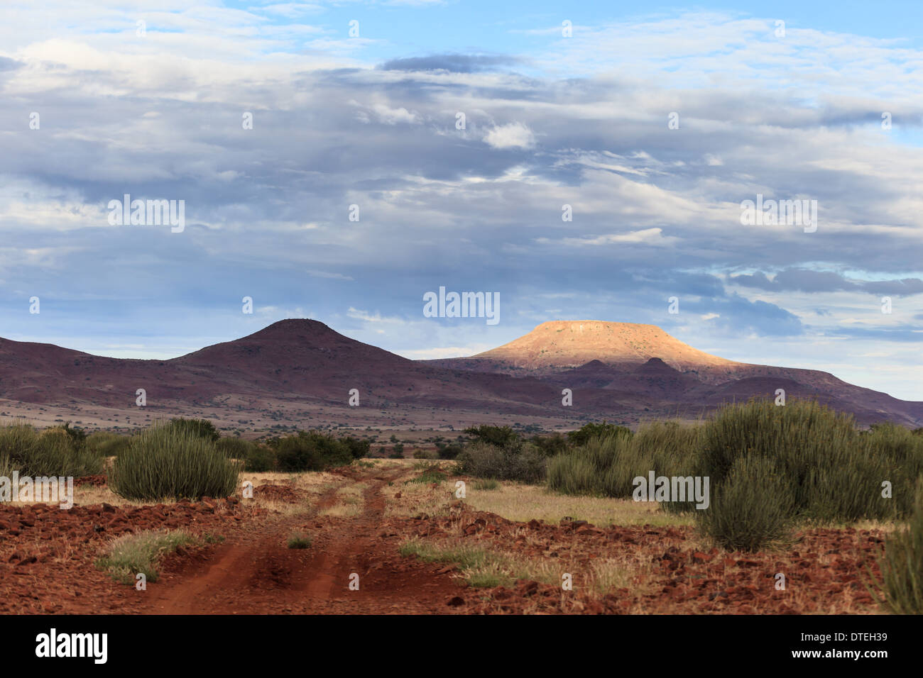 Unbefestigte afrikanische Straße überquert kleine Hügel, die in Richtung der Berge Namibias führen, von denen einer hell in der untergehenden Sonne leuchtet. Stockfoto