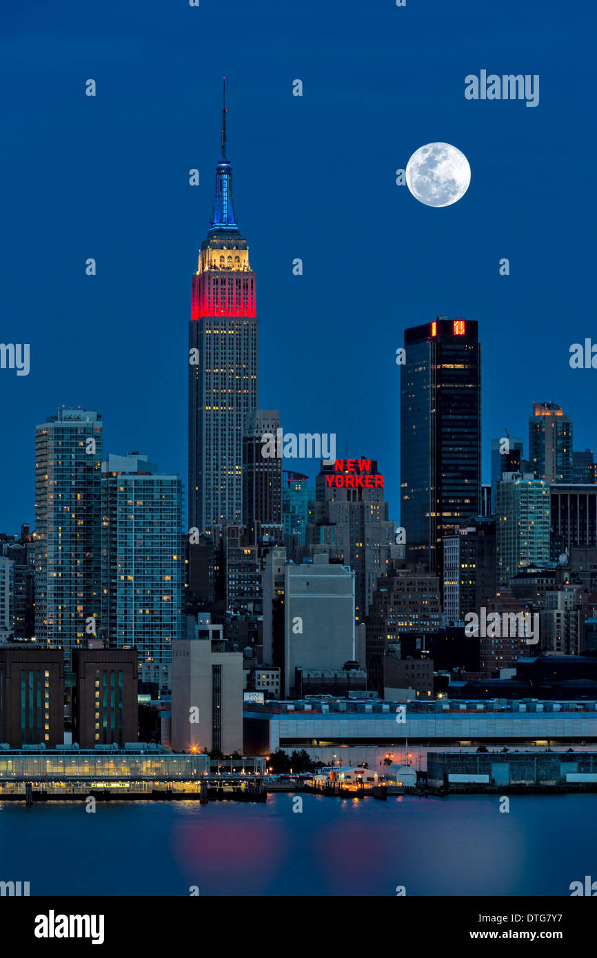 Der Vollmond steigt über die Skyline von New York City (NYC) während die Dämmerstunde. Ein Blick aus New Jersey über den Hudson River mit dem Empire State Building in rot, weiß und blau beleuchtet. Stockfoto
