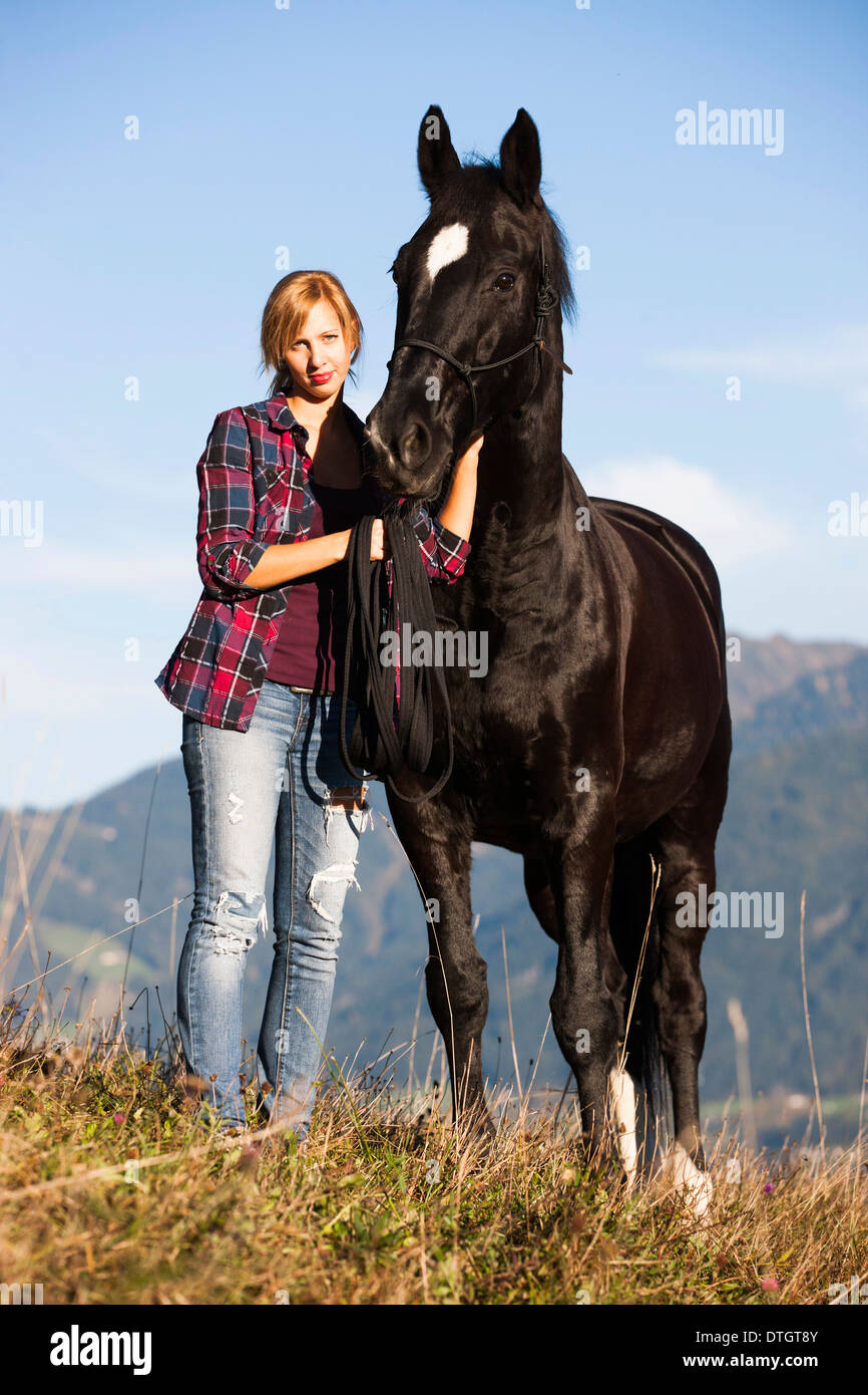 Eine junge Frau und ein schwarzes Hannoveraner Pferd stehend auf einer Bergwiese im Herbst, Nord-Tirol, Österreich Stockfoto