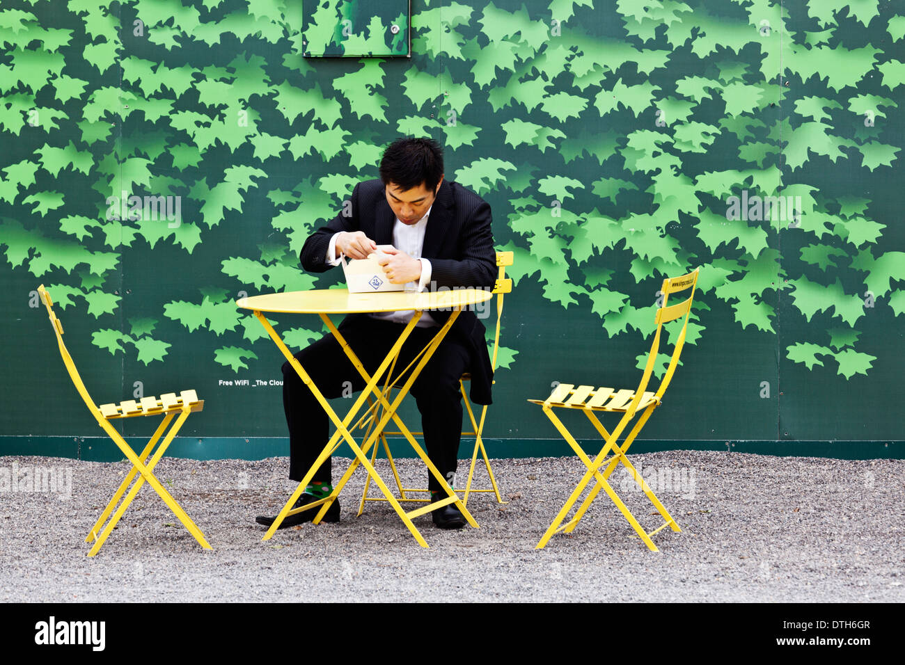 Ein asiatischer Mann in einem Anzug isst zum Mitnehmen an einem gelben Tisch und Stühle vor architektonischen Werbetafeln. Stockfoto