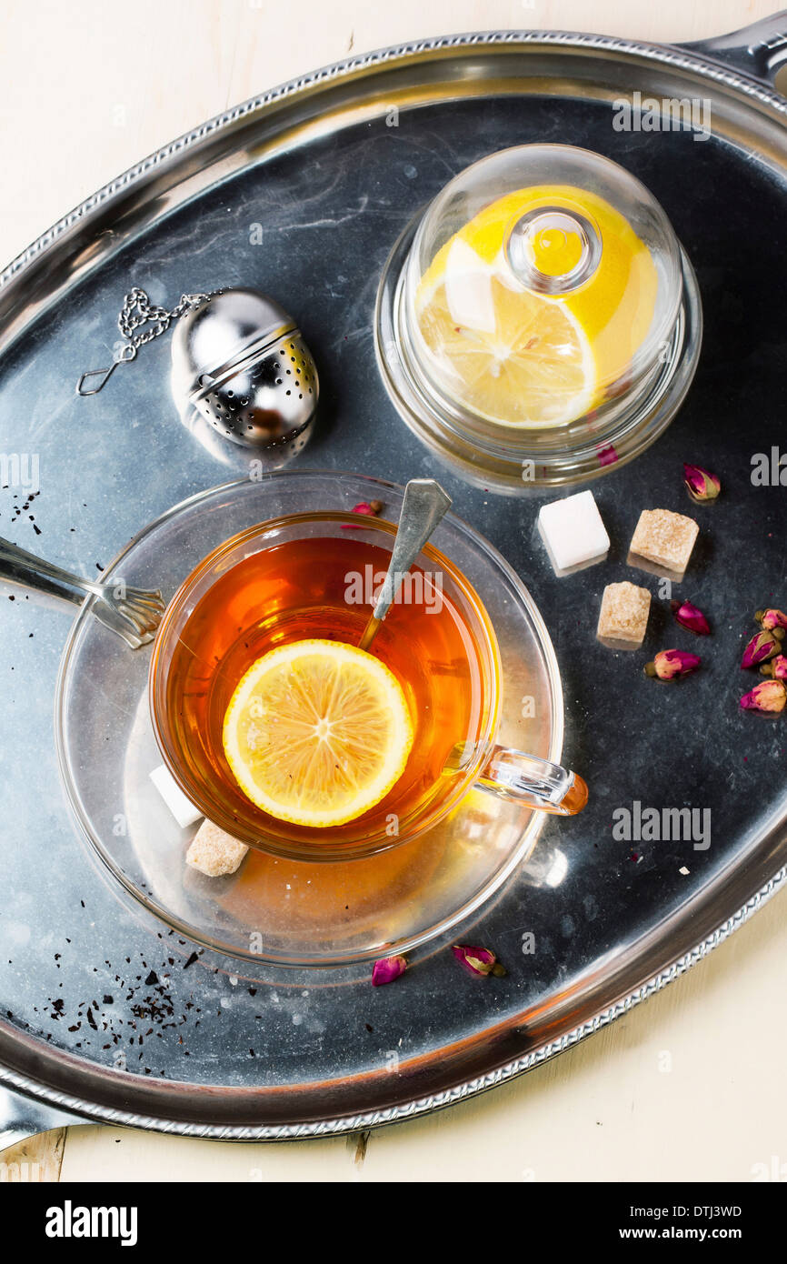 Draufsicht auf Glastasse Tee mit Würfelzucker, Zitrone und trockenen Rosenknospen, serviert auf Silber Vintage Tablett auf weißer Holztisch. Stockfoto