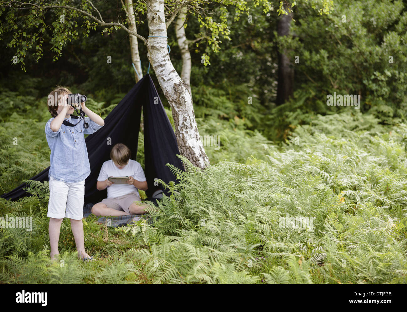 Zwei jungen camping in eine neue Gesamtstruktur sitzen unter einem schwarzen Leinwand Tierheim ein Junge Blick durch ein Fernglas Hampshire England Stockfoto