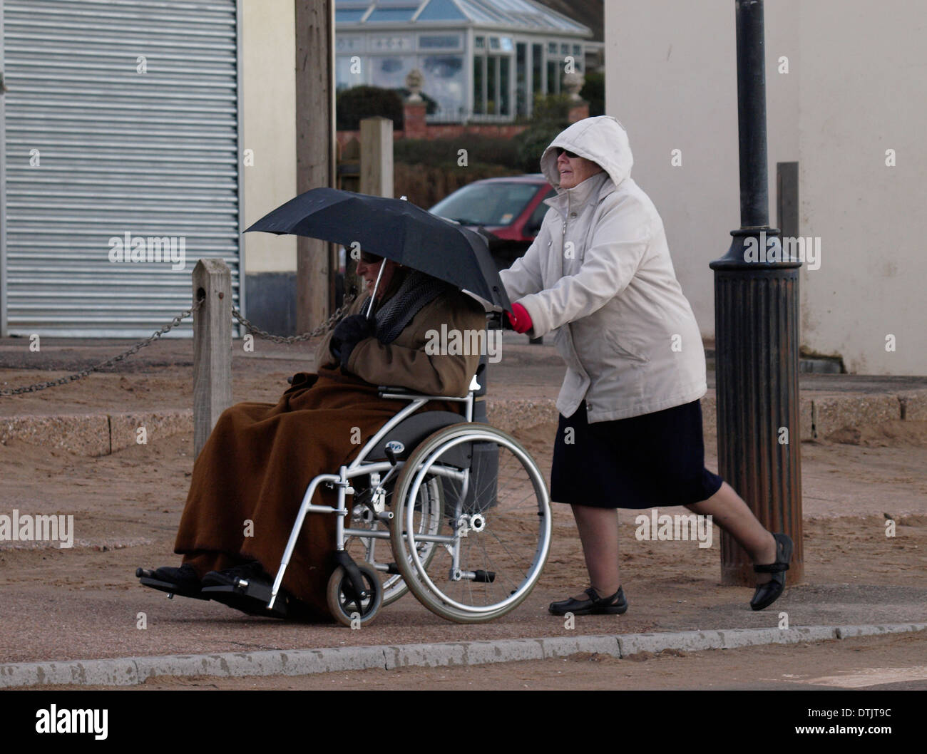 Alte Frau schieben Alter Mann im Rollstuhl hält Regenschirm im Winter, Exmouth, Devon, UK Stockfoto